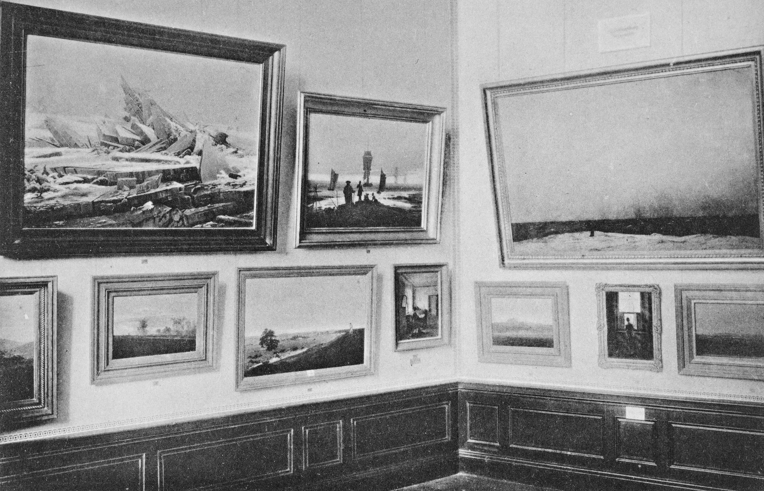 View of a gallery with works by Caspar David Friedrich at the German Centenary Exhibition of 1906 showing works hung on white walls in two rows, one above the other. Recognizable here, among others, are “The Sea of Ice,” the “Stages of Life,” and “Monk by the Sea.”